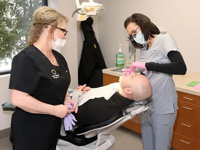 Dentist in black scrubs examining patient in dental chair while assistant observes in clinical office setting.