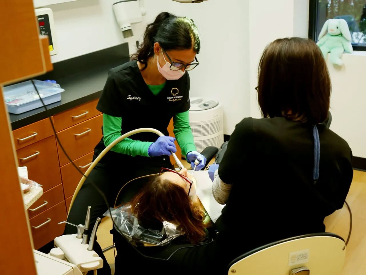 Dentist examining patient's teeth in dental office treatment room with assistant present.