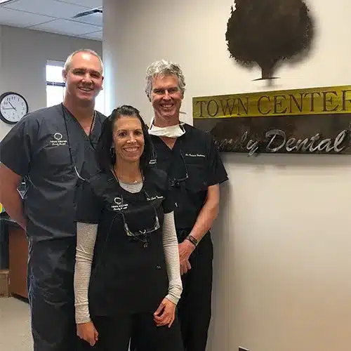 Three dental professionals in scrubs pose together in a modern dental office hallway.