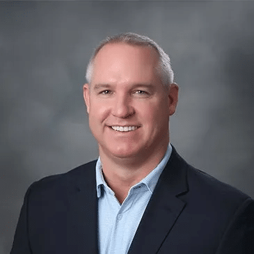 Professional headshot of smiling man in business casual attire against gray background.