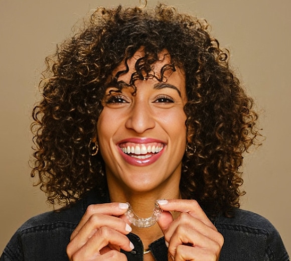 Smiling woman with curly hair wearing denim jacket against tan background, holding necklace.