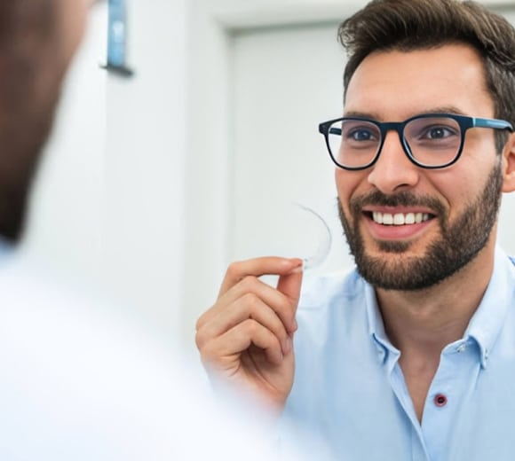 Smiling male dentist wearing glasses holding dental model in clinical office setting.