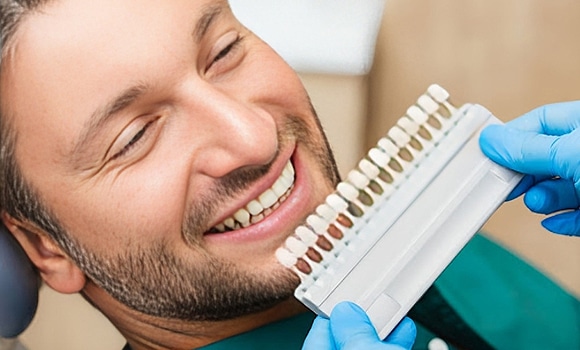 Dentist holding tooth shade guide next to smiling patient's face in dental office.