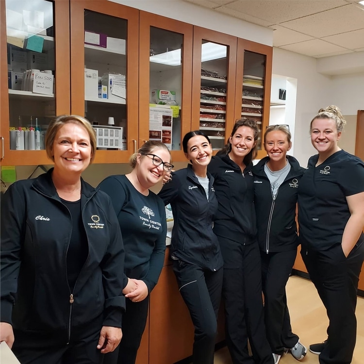 Six dental staff members in black uniforms smiling together in a modern dental office.