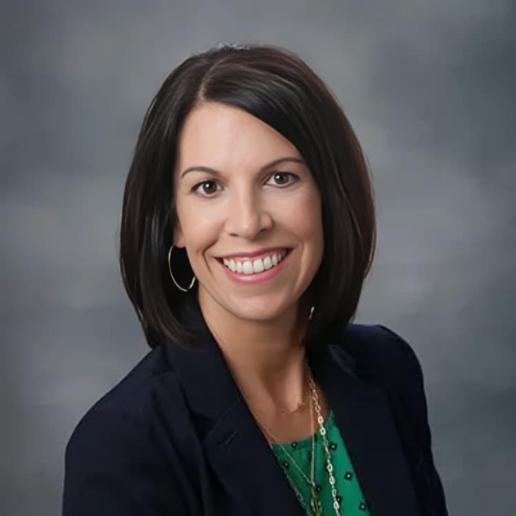 Professional headshot of smiling woman in dark blazer and green top against gray background.