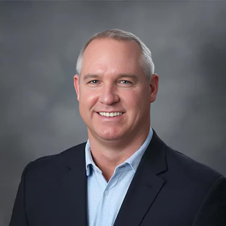 Professional headshot of smiling male dentist in dark blazer and light blue shirt against gray background.