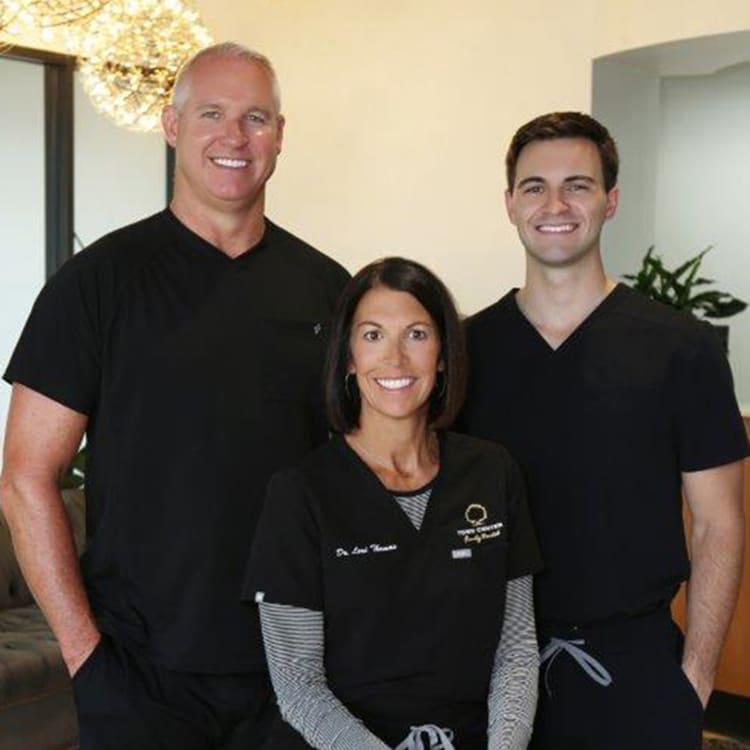 Three dental professionals in black uniforms smiling at camera in modern dental office setting.
