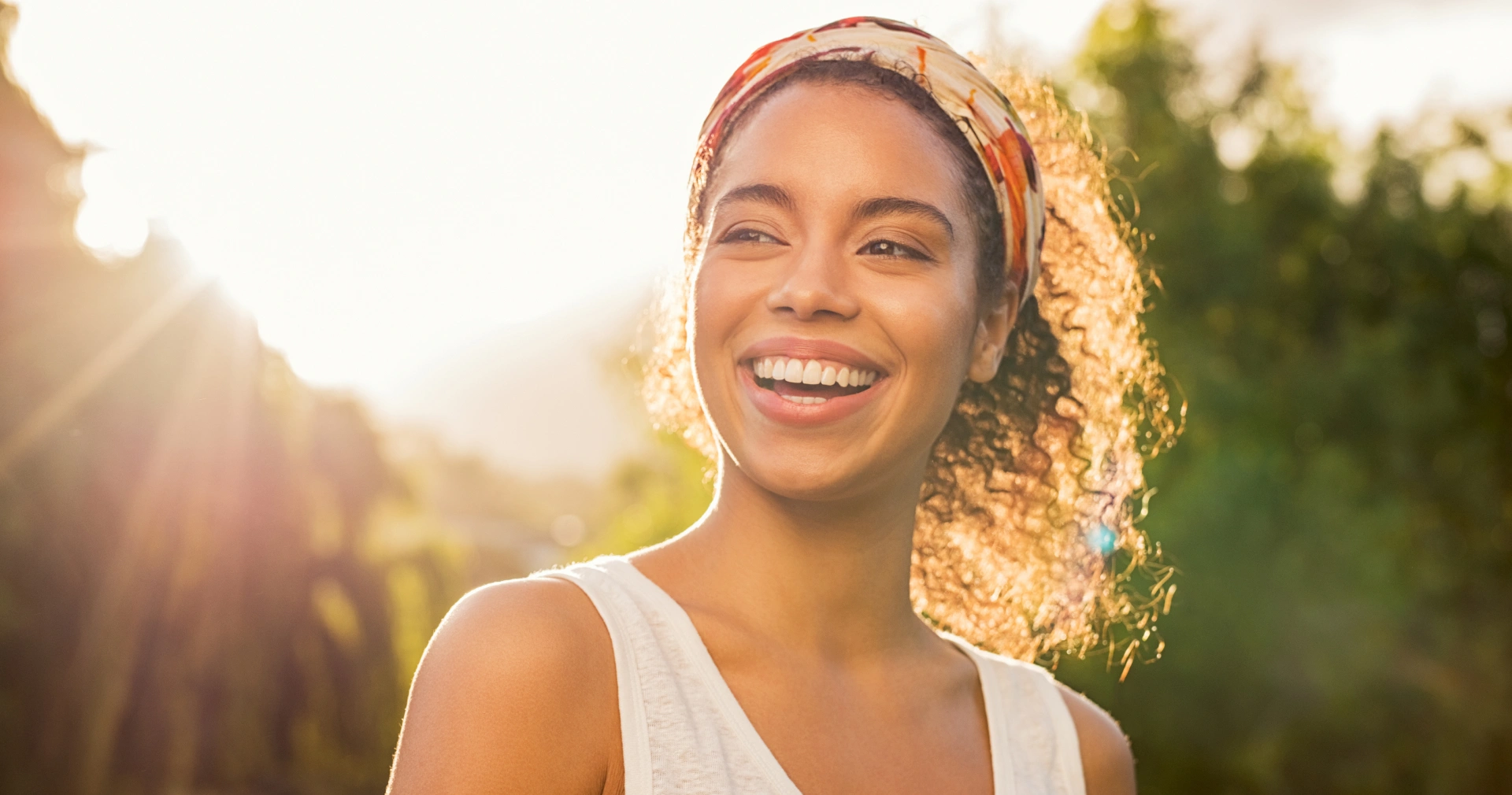 Smiling woman with curly hair wearing sunglasses outdoors in sunny, natural setting with green background.