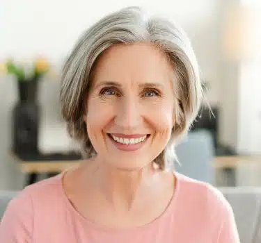 Professional headshot of smiling woman with gray hair wearing pink top in dental office setting.