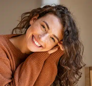 Smiling woman in brown sweater tilts head toward camera in warm, neutral indoor setting.