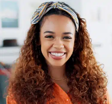 Smiling woman with curly reddish-brown hair wearing orange in bright dental office setting.