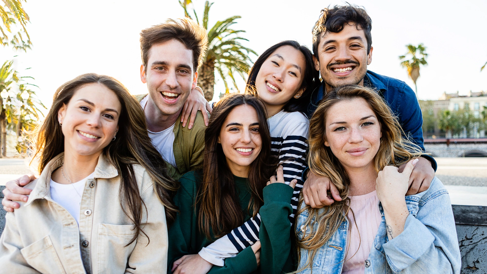 Group of six smiling people posing together outdoors with palm trees in background.