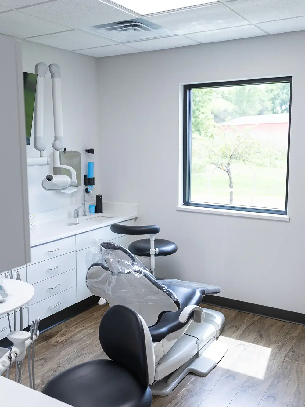 Modern dental examination room with black and white patient chair, white cabinetry, and large window in Flint, MI.