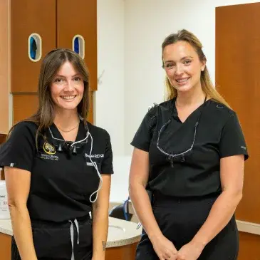 Two dental professionals in black scrubs smile in a modern dental office treatment room.