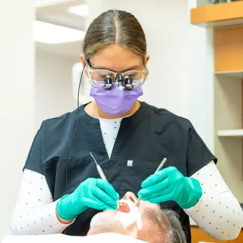 Dentist in purple mask and protective eyewear examining patient's teeth during preventative care visit.