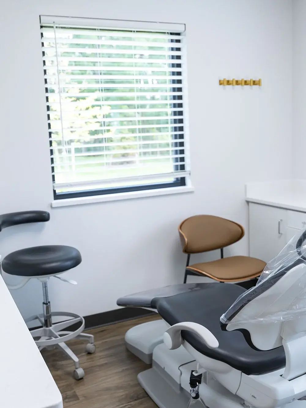 Modern dental treatment room with white chair, operator stool, patient seating, and window blinds in Flint.